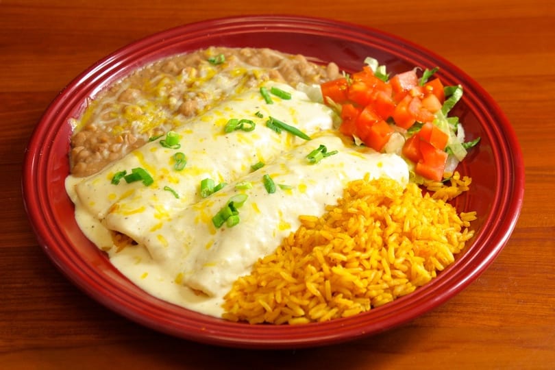 Two enchiladas with white sauce, rice, refried beans, and a side salad on a red plate.