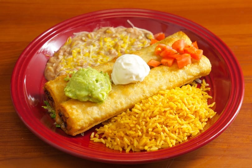 Two golden-fried flautas topped with guacamole, sour cream, and diced tomatoes, served with rice and refried beans on a red plate.