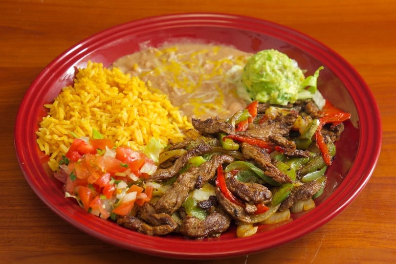 A red plate holds steak fajitas with peppers and onions, yellow rice, refried beans, pico de gallo, and guacamole.