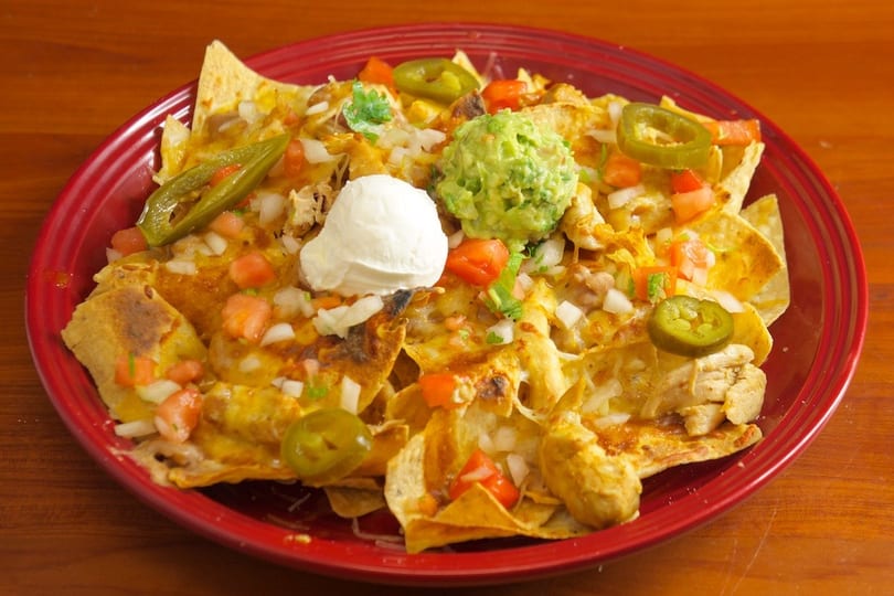 Chicken nachos with melted cheese, diced tomatoes, onions, jalapeños, guacamole, and sour cream on a red plate.